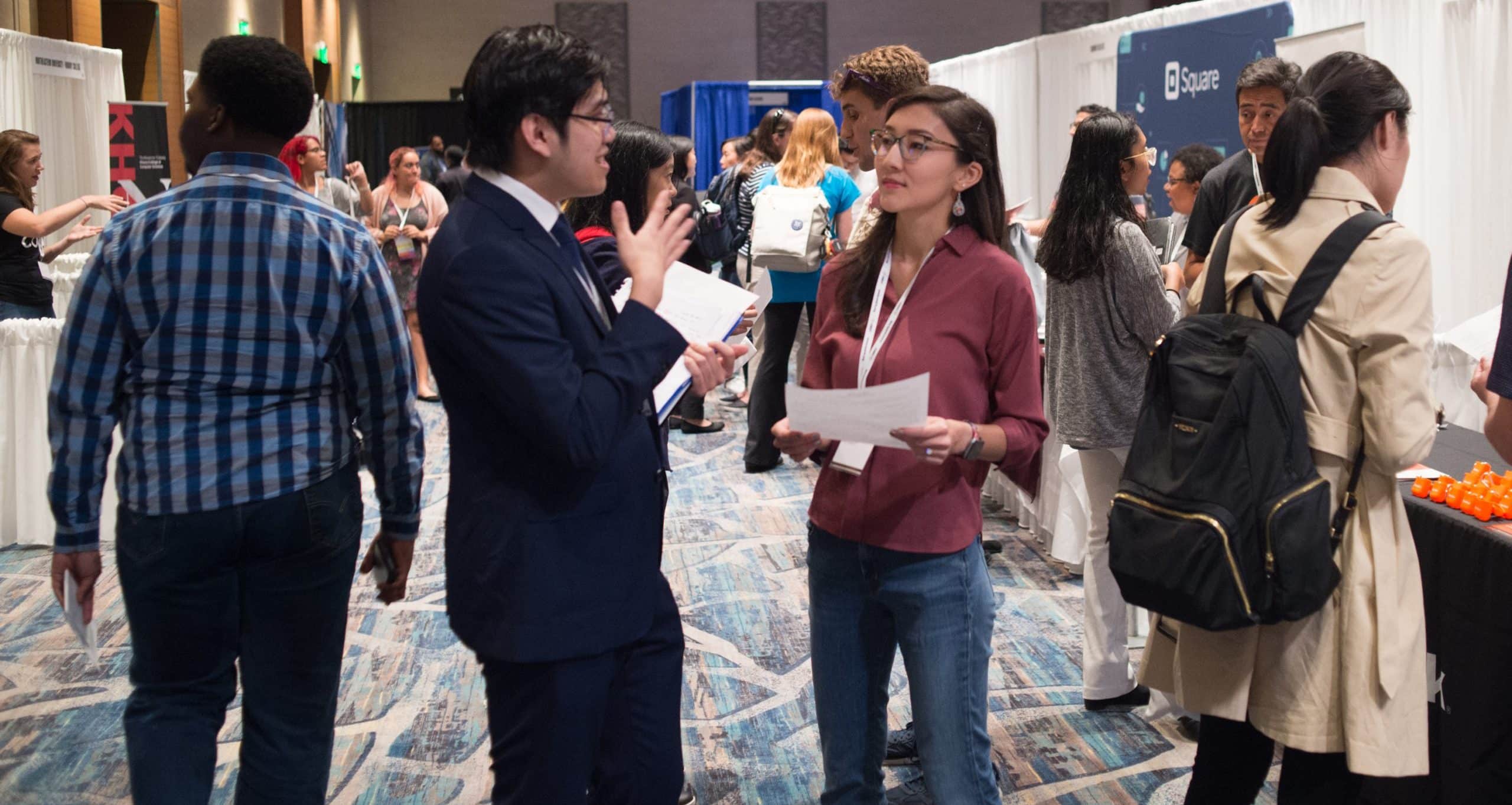 At a bustling business event, people in professional attire engage in conversations, some holding papers. Booths and representatives offer information on various initiatives, including programs for minorities, fostering an inclusive environment.